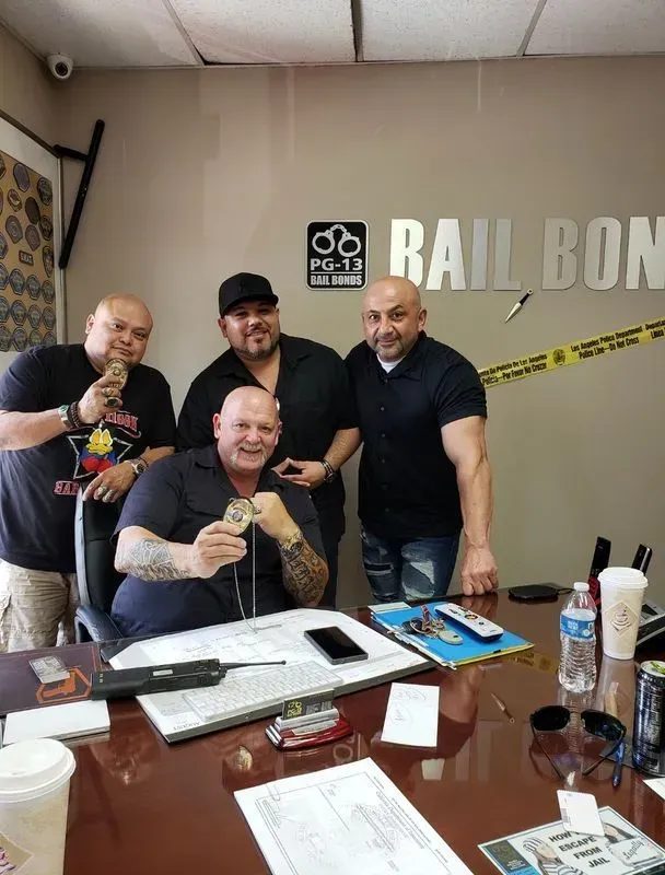 Four men pose in a office, one seated at a desk holding a trophy, with a “Bail Bond” sign behind them.